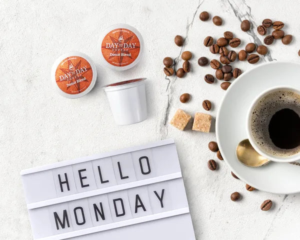 Flat lay of Day to Day Coffee Donut Blend K-Cup pods with coffee beans and Hello Monday sign on white marble
